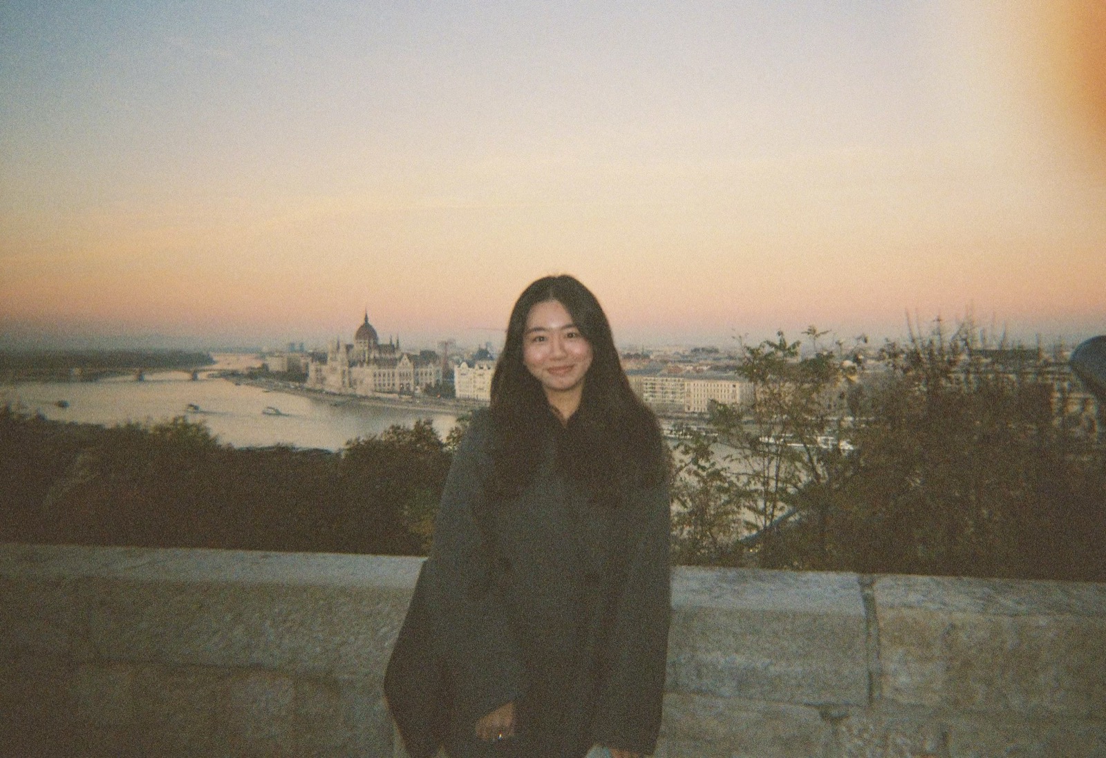 Joyce standing in front of the Danube River and Fisherman's Bastion in Budapest, Hungary at sunset, wearing a grey coat