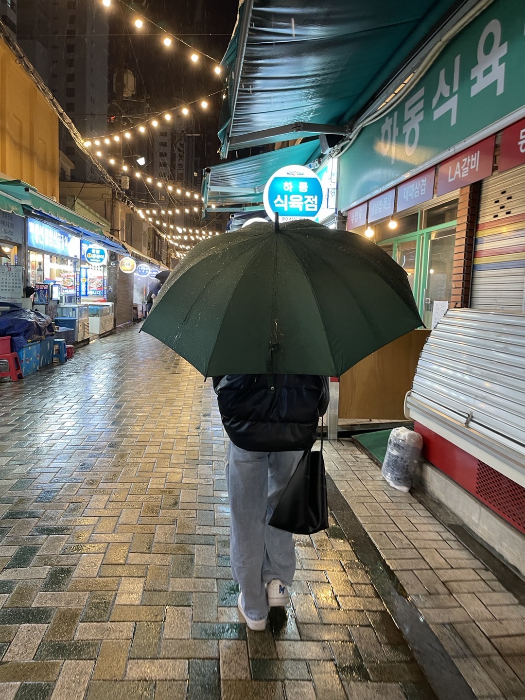 Joyce walking in the Haeundae Traditional Market with a black puffer jacket, blue jeans, and umbrella