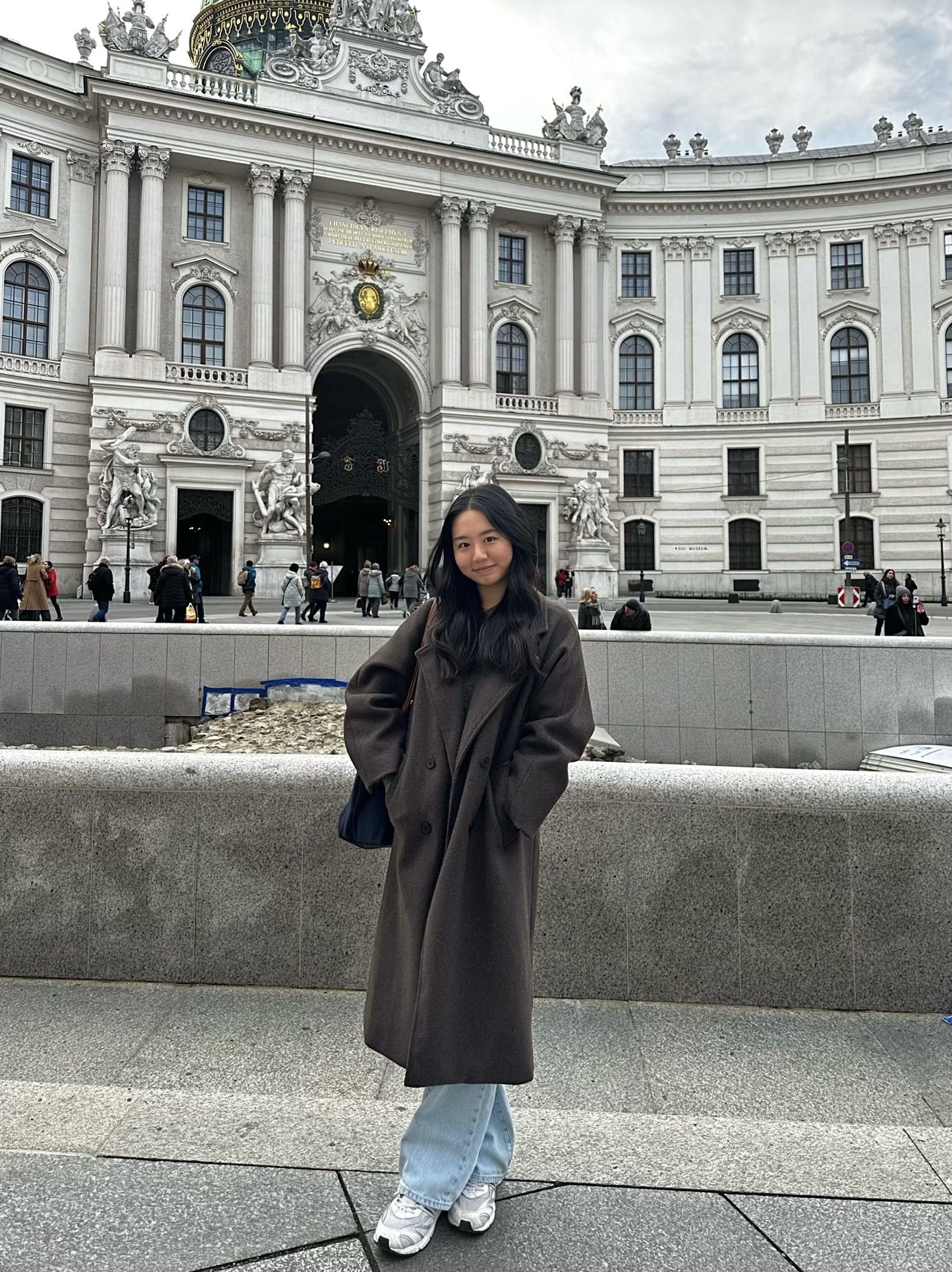 Joyce standing in front of the Hofburg Palace in Vienna in a brown coat
